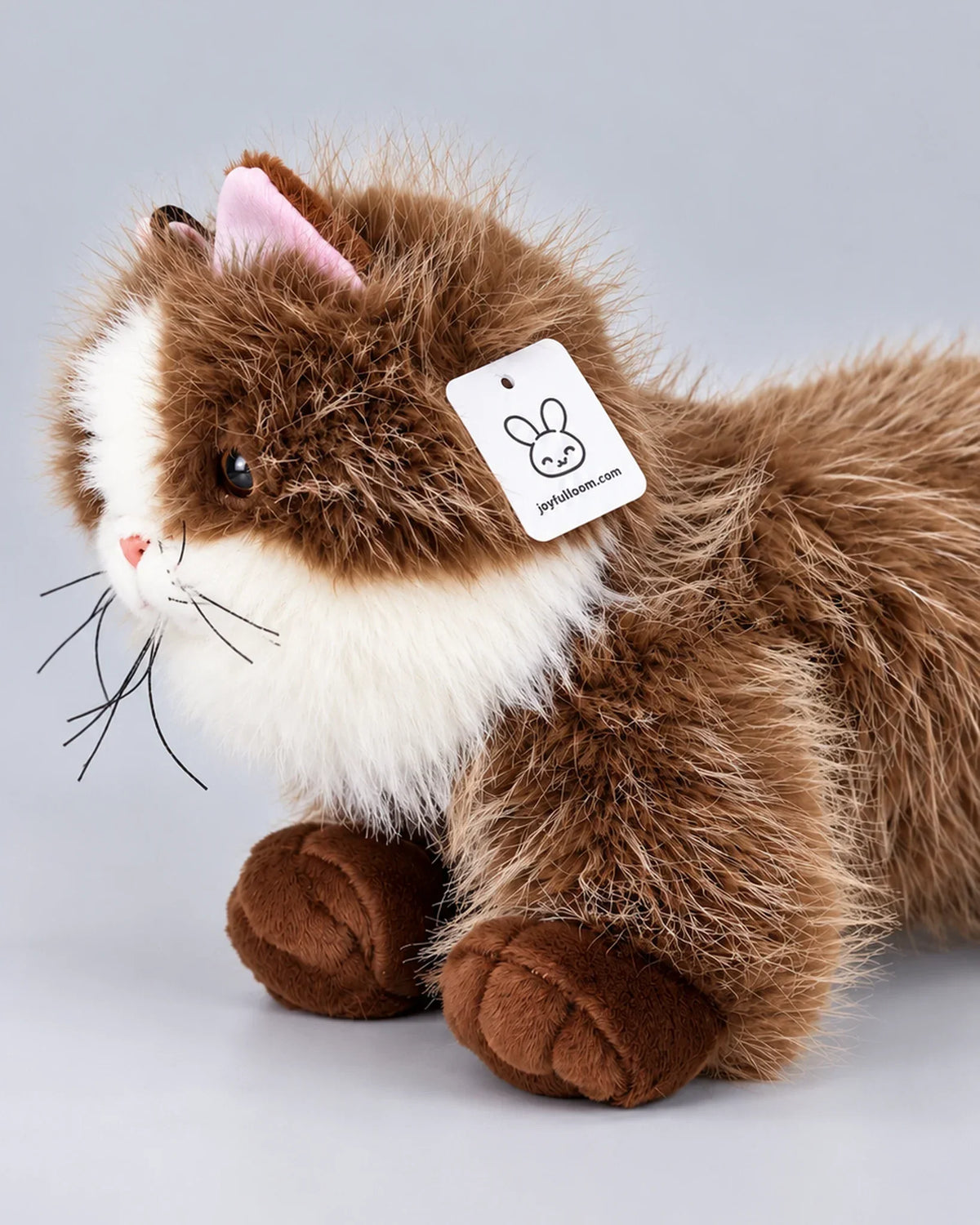 Realistic brown and white Ragdoll-style cat plush toy, lying down, studio photo